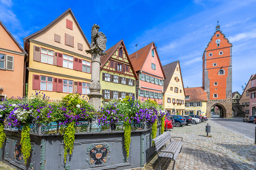 Bunte Häuser und Fachwerk sowie ein Tor mit roter Fassade säumen eine Kopfsteinpflasterstraße in Dinkelsbühl. Blumen blühen am Rand eines Brunnens. Dinkelsbuehl Deutschland Innenstadt