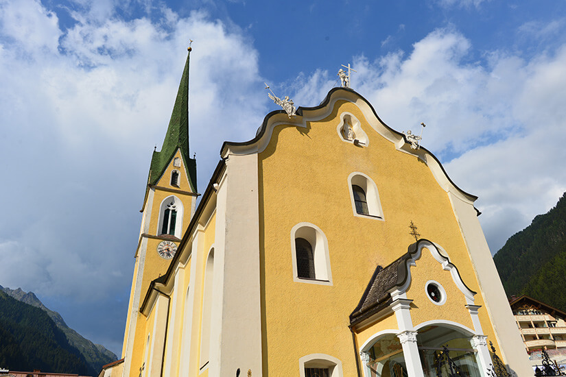 Zu erkennen ist eine gelb-weiße Kirche mit geschwungener Barockfassade und einem hohen, spitzen Kirchturm. Im Hintergrund erheben sich bewaldete und felsige Alpenberge unter einem blau bewölkten Himmel. Ischgl Kirche