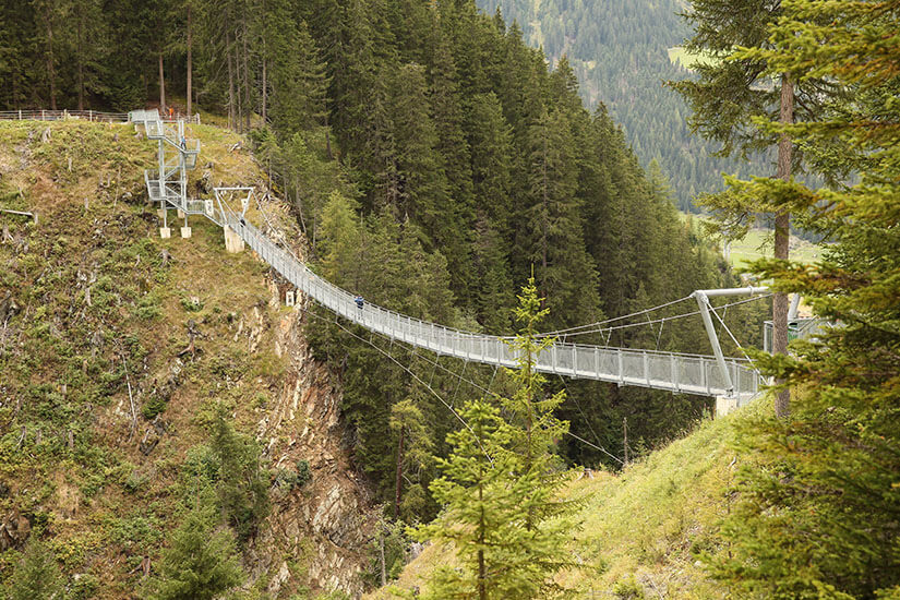 Zu erkennen ist eine lange Hängebrücke, die eine tiefe, bewaldete Schlucht in einer alpinen Landschaft überspannt. Rundherum wachsen dichte Nadelwälder auf den steilen Berghängen. Tirol Haengebruecke