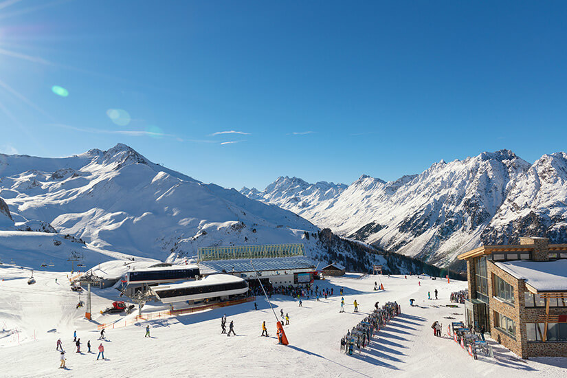 Zu erkennen ist ein weitläufiges Skigebiet mit zahlreichen Wintersportlern, Liftanlagen und Gebäuden inmitten verschneiter Berge. Die schroffen, schneebedeckten Gipfel bilden eine eindrucksvolle alpine Kulisse unter klarem blauem Himmel. Ischgl Skifahren