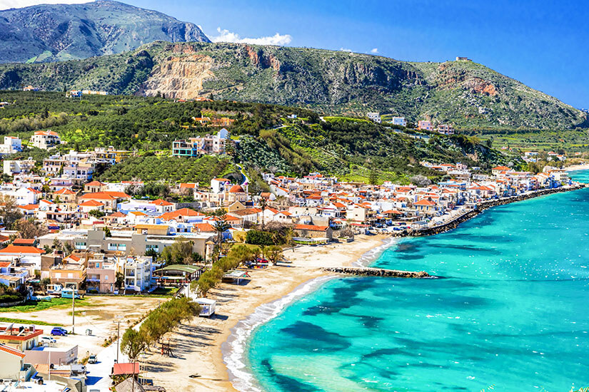 Ein Küstenort auf der Insel Kreta ist zu sehen. Das Wasser leuchtet türkisblau. Weiße Häuser mit roten Dächern stehen am Ufer, im Hintergrund zeigt sich grüne Natur. All Inclusive unter 1000 Euro Kreta