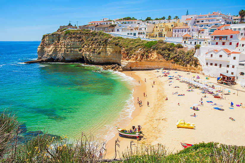 Eine bunte Küstenstadt thront über einer Klippe am Meer. Heller Sandstrand und grünblaues Meer sind zu sehen. All Inclusive unter 1000 Euro Algarve