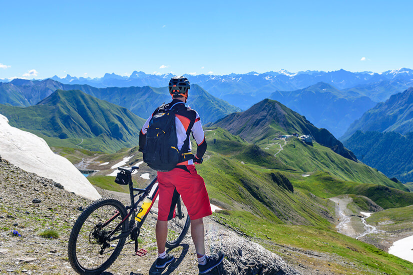 Zu erkennen ist ein Mountainbiker mit Fahrrad auf einem hoch gelegenen Bergpfad. Vor ihm erstreckt sich ein beeindruckendes Panorama aus grünen Bergrücken, tiefen Tälern und fernen Alpengipfeln. Ischgl Mountainbike
