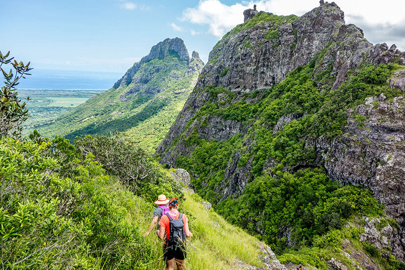 Auf einem schmalen, grasbewachsenen Pfad wandern zwei Personen mit Rucksäcken und Sonnenhut durch die üppig grüne Berglandschaft von Mauritius. Packliste Mauritius Wanderung