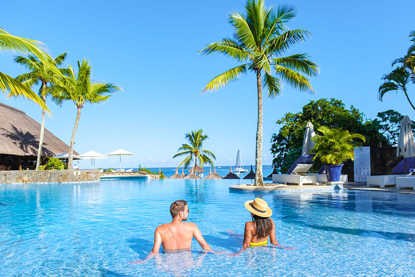 Im türkisblauen Infinity-Pool eines Resorts auf Mauritius entspannt ein Paar, die Frau mit gelbem Bikini und Strohhut, und blickt auf das Meer hinaus. Packliste Mauritius Pool