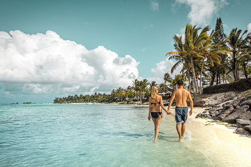 Hand in Hand schlendert ein Paar in Badekleidung durch das seichte, türkisfarbene Wasser entlang eines weißen Sandstrands auf Mauritius. Packliste Mauritius Strand