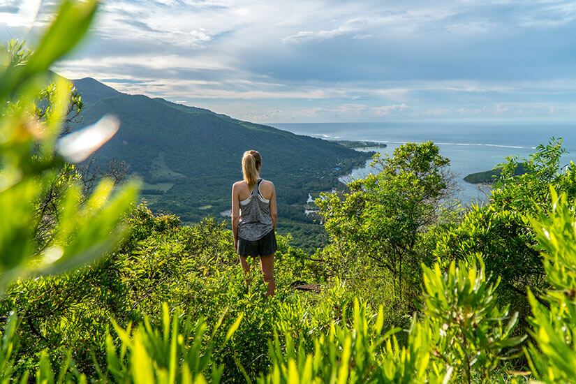 Inmitten üppiger, grüner Vegetation steht eine blonde Frau in Sportkleidung auf einer Anhöhe und genießt den weiten Ausblick über die Küste von Mauritius. Packliste Mauritius Ausblick