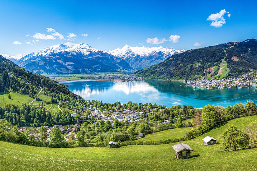 Zu erkennen ist ein idyllischer Alpensee, umgeben von grünen Wiesen, bewaldeten Berghängen und kleinen Ortschaften. Im Hintergrund erheben sich majestätische, teilweise schneebedeckte Berggipfel unter einem strahlend blauen Himmel. Oesterreich Land