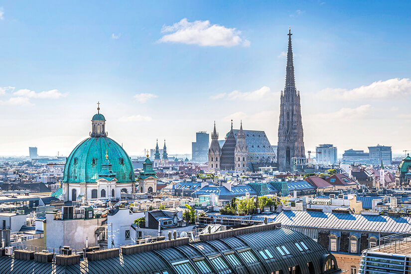 Zu erkennen ist die Wiener Innenstadt mit dem markanten Stephansdom und seinem hohen Südturm. Links fällt die große türkisfarbene Kuppel der Peterskirche ins Auge, während sich im Vordergrund zahlreiche Dächer der Altstadt erstrecken. Oesterreich Wien