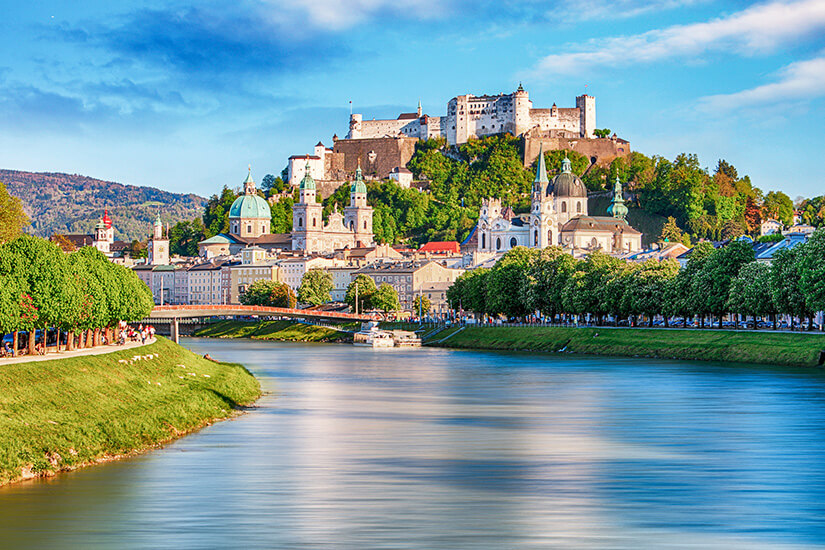 Zu erkennen ist Salzburg mit der Salzach im Vordergrund und der historischen Altstadt entlang des Ufers. Hoch über der Stadt thront die Festung Hohensalzburg, umgeben von markanten Kirchtürmen und Kuppeln. Oesterreich Salzburg