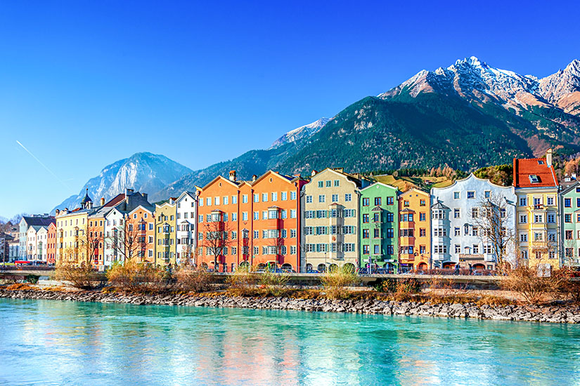 Zu erkennen ist Innsbruck mit einer Reihe farbenfroher Häuser direkt am Ufer des Inns. Im Hintergrund erheben sich eindrucksvolle, teilweise schneebedeckte Gipfel der Alpen. Oesterreich Innsbruck