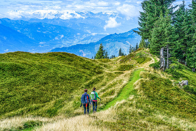 Zwei Wanderer sind auf einem schmalen Bergpfad durch eine grüne alpine Landschaft unterwegs. Im Hintergrund erstreckt sich ein beeindruckendes Panorama aus Tälern und teils schneebedeckten Berggipfeln. Oesterreich Wandern