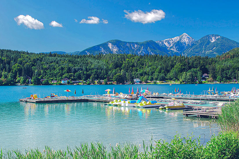 Zu erkennen ist ein klarer Alpensee mit zahlreichen Tret- und Freizeitbooten an einem Steg. Im Hintergrund erstrecken sich bewaldete Ufer und markante Berggipfel unter blauem Himmel. Oesterreich See