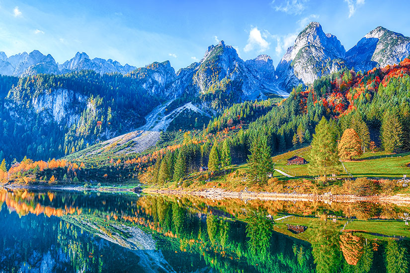 Zu erkennen ist eine eindrucksvolle Berglandschaft im Herbst mit schroffen Gipfeln, bunten Wäldern und grünen Wiesen. Ein ruhiger See im Vordergrund spiegelt die Berge und die farbenprächtige Vegetation nahezu vollständig wider. Oesterreich Herbst