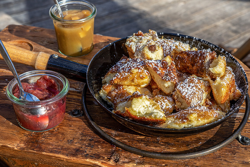 Zu erkennen ist Kaiserschmarrn, mit Puderzucker bestreut und in einer kleinen Pfanne serviert. Dazu stehen zwei Gläser mit Fruchtkompott beziehungsweise Apfelmus bereit. Oesterreich Essen