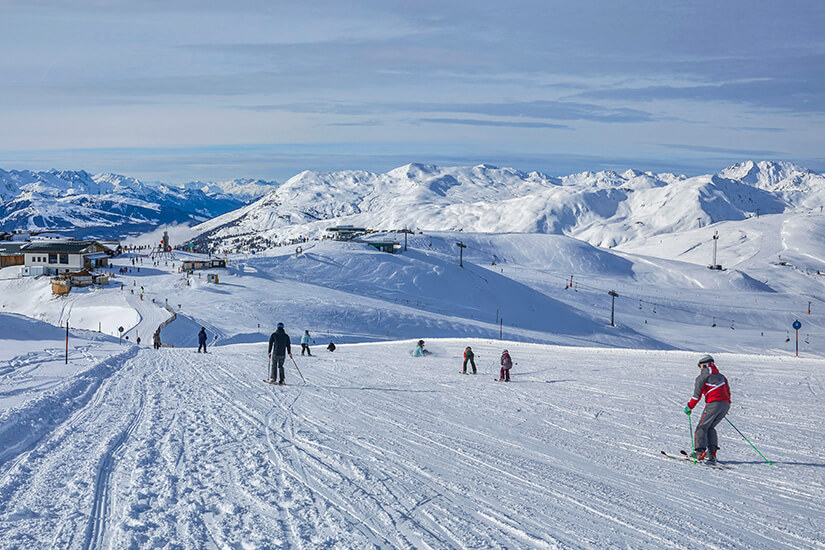 Zu erkennen ist ein weitläufiges Skigebiet mit mehreren Skifahrern auf einer breiten, verschneiten Piste. Im Hintergrund erstreckt sich ein beeindruckendes Panorama schneebedeckter Berggipfel mit Liftanlagen. Oesterreich Skigebiet