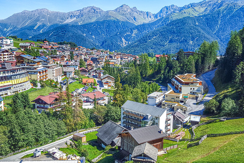 Zu erkennen ist der österreichische Alpenort Serfaus mit zahlreichen traditionellen Häusern, Hotels und grünen Wiesen. Im Hintergrund erheben sich imposante Berggipfel, deren Hänge teilweise dicht bewaldet sind. Tirol Serfaus