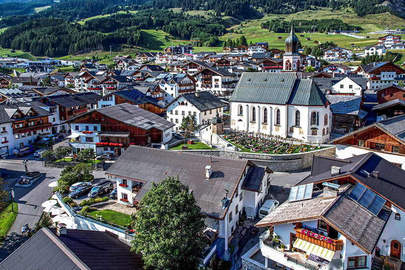 Zu erkennen ist der malerische Tiroler Ort Fiss mit zahlreichen traditionellen Alpenhäusern und einer markanten Kirche im Ortszentrum. Rund um die Kirche liegt ein gepflegter Friedhof, während sich im Hintergrund grüne Wiesen und bewaldete Berghänge erstrecken. Tirol Fiss