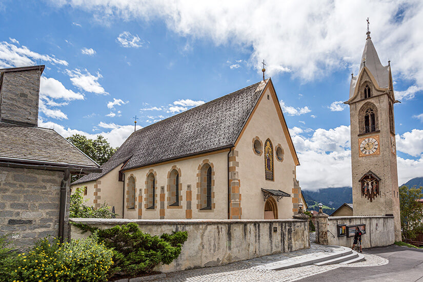 Zu erkennen ist eine historische Kirche mit hell verputzten Mauern, hohen Rundbogenfenstern und einem steilen Schindeldach. Daneben steht ein freistehender Glockenturm mit einer großen Uhr und einem spitzen Turmhelm. Serfaus-Fiss-Ladis Kirche