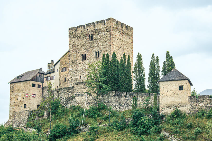 Zu erkennen ist eine mittelalterliche Burganlage aus Naturstein mit einem mächtigen, zinnenbekrönten Wehrturm. Die historischen Gebäude sind von einer hohen Burgmauer, schlanken Bäumen und dichter grüner Vegetation umgeben. Serfaus-Fiss-Ladis Burg