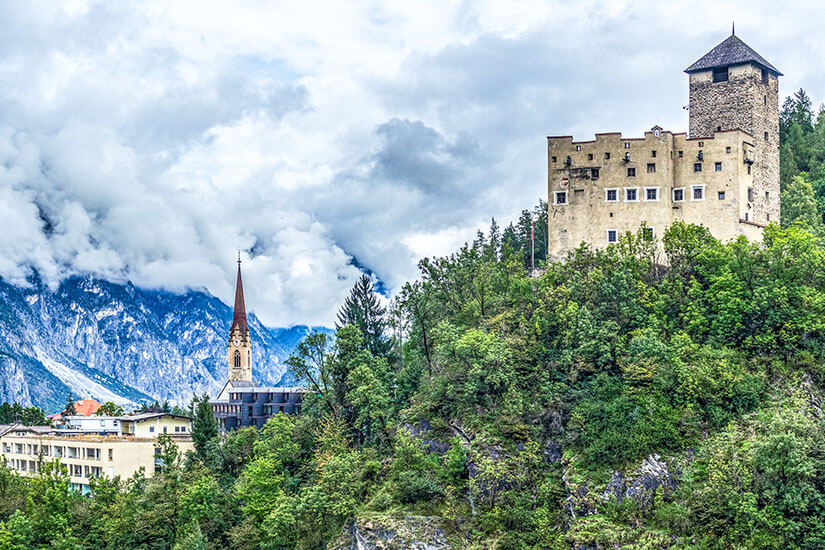 Zu erkennen ist eine imposante mittelalterliche Burg mit einem hohen, quadratischen Turm, die auf einem bewaldeten Hügel thront. Links davon ragt ein schlanker Kirchturm empor, während sich im Hintergrund mächtige, teilweise von Wolken verhüllte Alpenberge erheben. Oesterreich Schloss