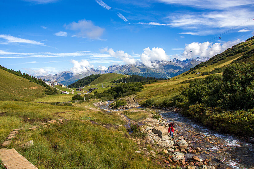 Zu erkennen ist eine idyllische Alpenlandschaft mit grünen Bergwiesen, einem plätschernden Bach und einem Wanderweg. Eine Person steht am felsigen Bachufer, während sich im Hintergrund beeindruckende Berggipfel und eine Seilbahn unter einem blauen Himmel erstrecken. Tirol Wandern