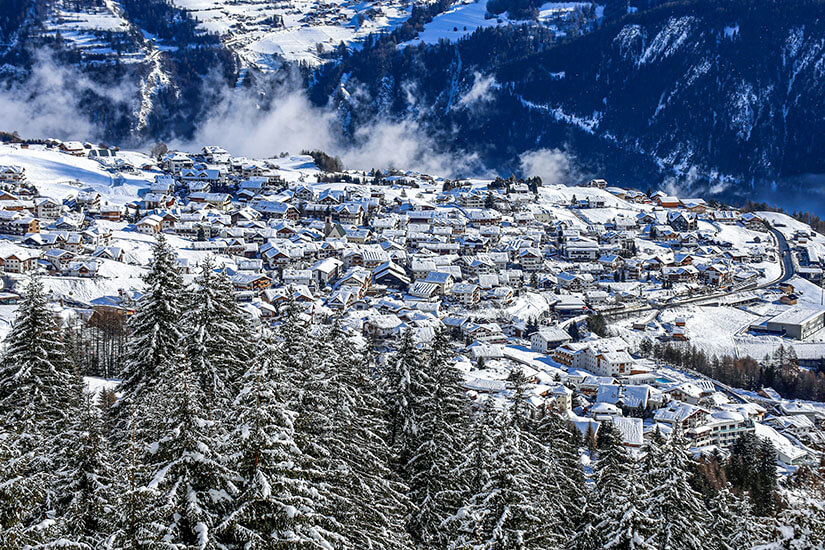 Zu erkennen ist ein verschneites Bergdorf in der Region Serfaus-Fiss-Ladis mit zahlreichen schneebedeckten Häusern. Im Vordergrund stehen dicht bewachsene, weiß verschneite Nadelbäume, während im Hintergrund bewaldete Berghänge und aufsteigende Nebelschwaden zu sehen sind. Serfaus-Fiss-Ladis Winter