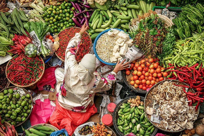 Von oben betrachtet sitzt eine Marktfrau mit hellem Kopftuch und geblümtem Gewand inmitten ihrer Körbe voller frischer Waren auf einem malaysischen Markt. Malaysisches Essen Markt