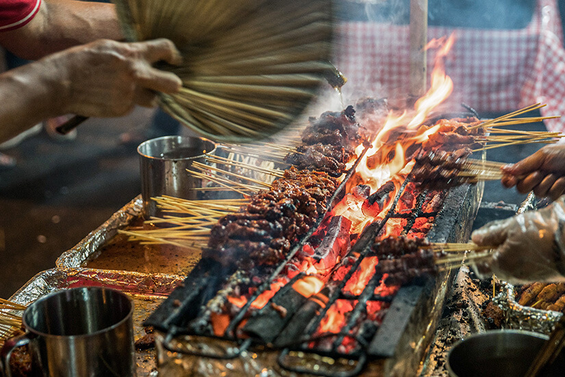 Über glühender Holzkohle brutzeln zahlreiche Satay-Spieße, während ein Straßenkoch mit einem geflochtenen Fächer die Flammen anfacht und eine weitere Hand mit Handschuh die Spieße wendet. Malaysisches Essen Street Food