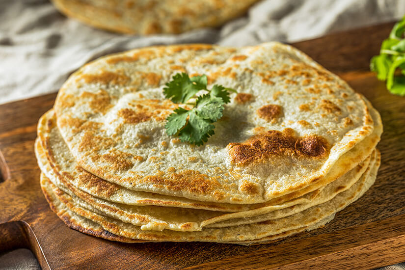 Auf einem dunklen Holzbrett stapeln sich mehrere dünne, goldbraun gebackene Fladenbrote mit knusprig gebräunten Stellen, garniert mit einem frischen Korianderzweig. Malaysisches Essen Roti Canai