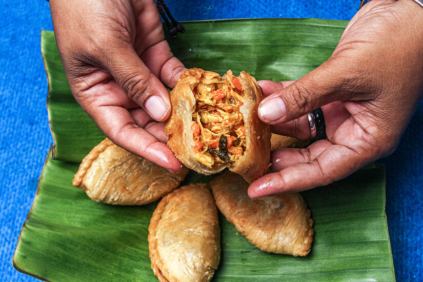 Zwei Hände brechen ein goldbraun frittiertes Karipap auf und geben den Blick auf die würzige Currfüllung mit Kartoffeln, Karotten und Curryblättern frei. Malaysisches Essen Karipap