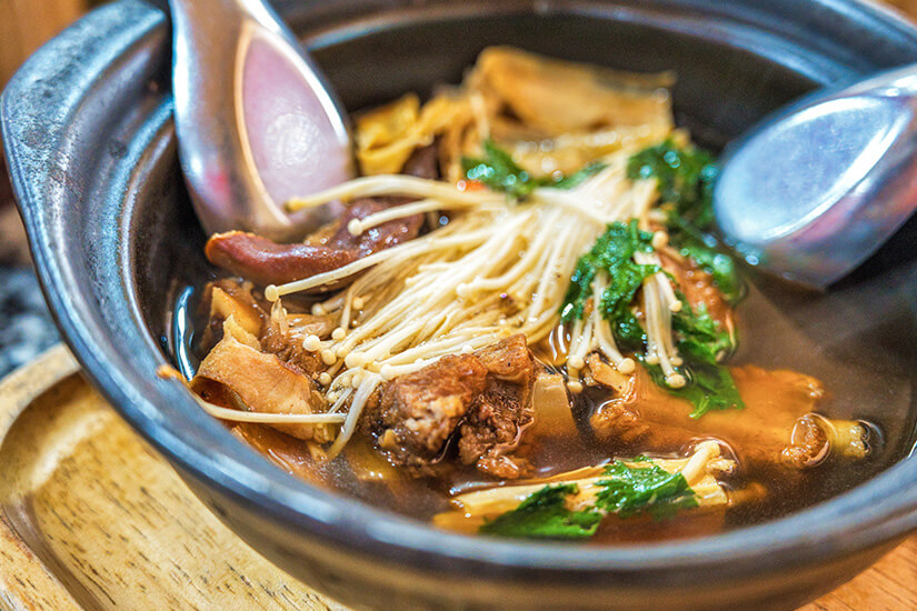 In einem dunklen Tontopf dampft Bak Kut Teh, eine kräftige malaysische Kräuterbrühe mit zarten Schweinerippchen, Enoki-Pilzen, frischem Grün und Tofuhaut. Malaysia Bak Kut Teh