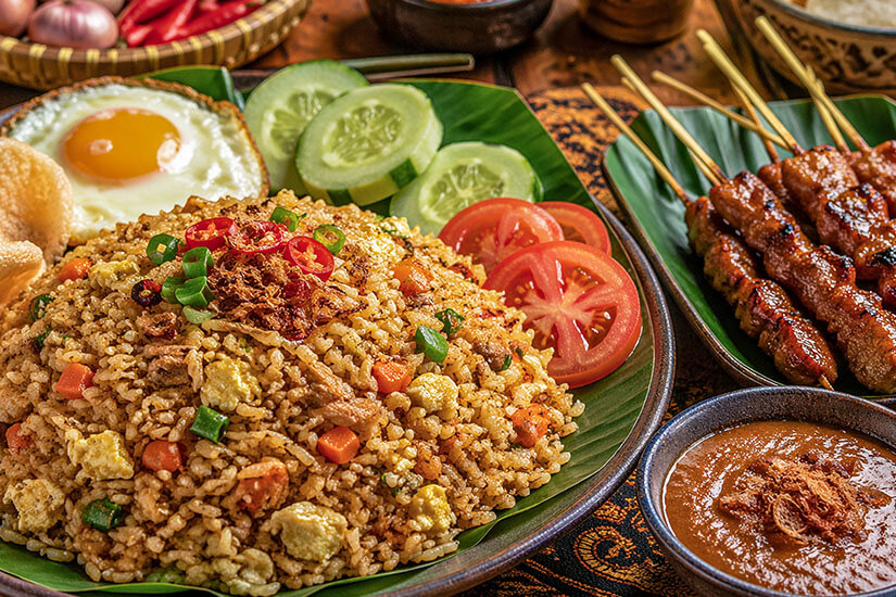 Auf einem Bananenblatt angerichtet, lockt eine üppige Portion Nasi Goreng mit Rührei, Karottenwürfeln, Frühlingszwiebeln und Chiliringen, begleitet von Spiegelei, Krabbenchips sowie Gurken- und Tomatenscheiben. Malaysisches Essen Nasi Goreng