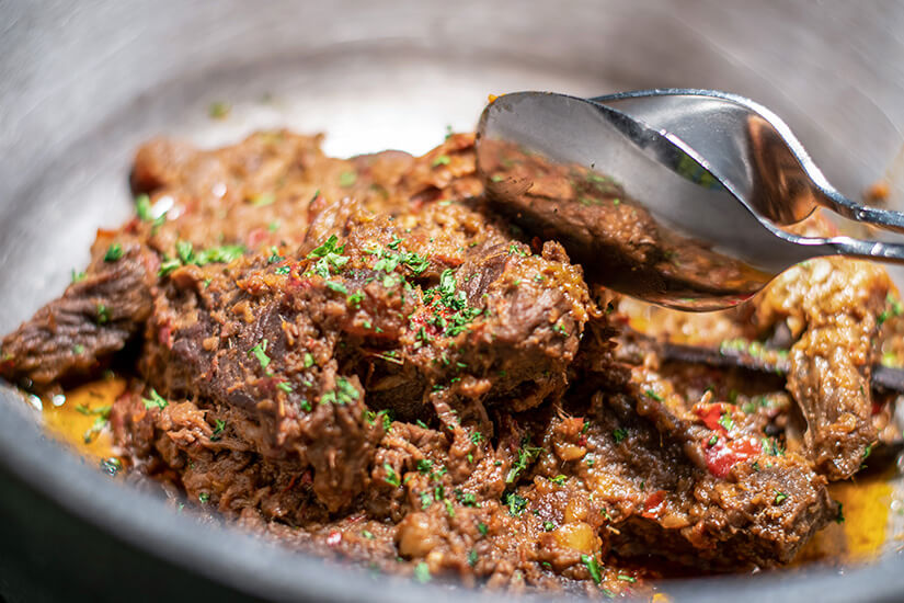 In einer Metallschale liegt dunkelbraunes, lange geschmortes Rindfleisch-Rendang, dessen zarte Fasern von einer dicken, würzigen Gewürzpaste überzogen und mit frisch gehackten Kräutern bestreut sind. Malaysisches Essen Rendang