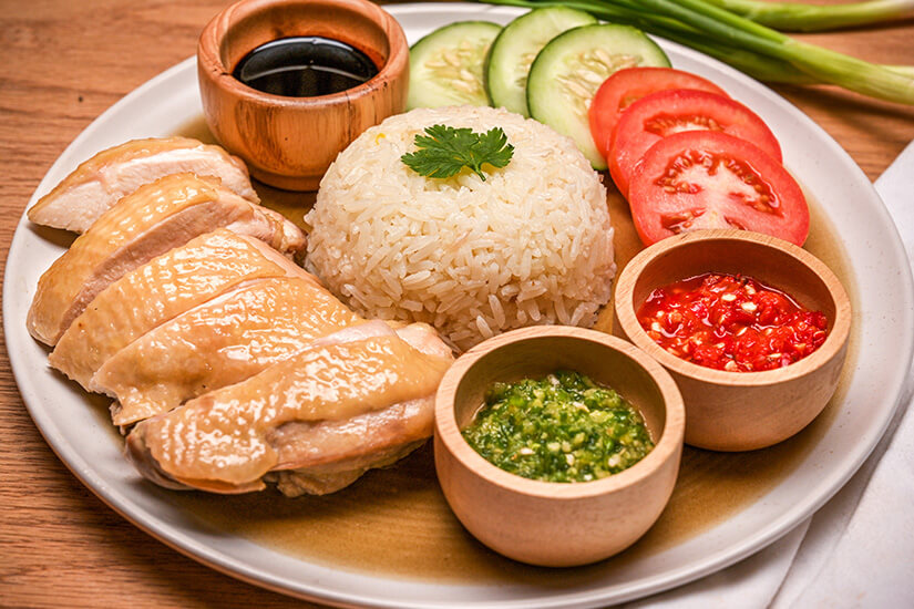 Auf einem weißen Teller ist Hainanese Chicken Rice angerichtet: zart pochierte Hähnchenstücke mit glänzender Haut neben einer Kuppel aus duftendem Reis, gekrönt von einem Korianderblatt. Hainanese Chicken Rice