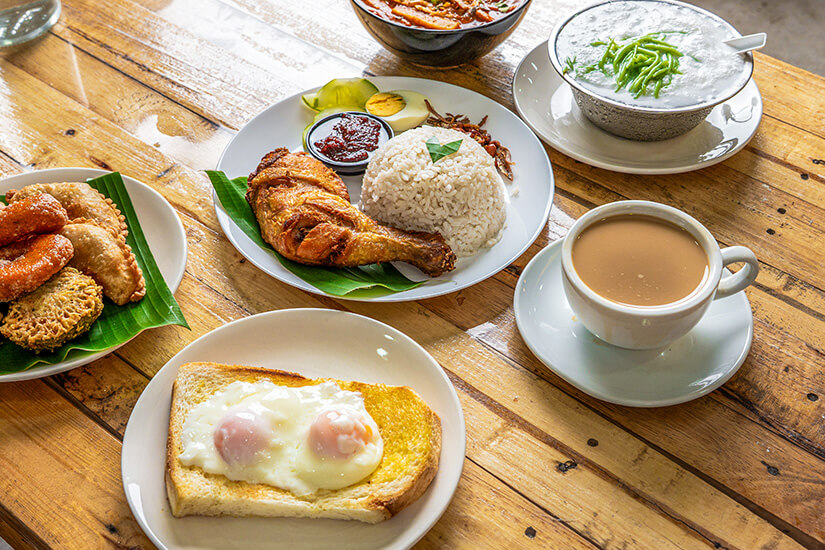 Auf einem rustikalen Holztisch ist ein malaysisches Frühstück angerichtet, mit Nasi Lemak samt knusprigem Brathähnchen, Sambal, Ei und Sardellen sowie einer Tasse milchigem Teh Tarik. Malaysisches Essen Tee
