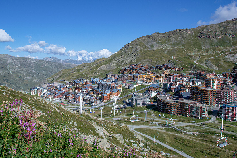 Panoramablick auf den hochgelegenen Wintersportort Val Thorens in den französischen Alpen im Sommer. Im Vordergrund blühen rosa Wildblumen am grünen Berghang, während im Hintergrund die schneefreien Gebirgskämme unter blauem Himmel mit Schönwetterwolken aufragen. Val Thorens Frankreich Sommer
