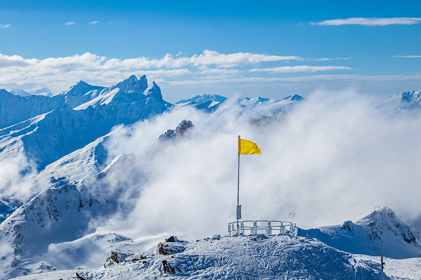 Aussichtsplattform auf dem verschneiten Gipfel der Cime de Caron in Val Thorens mit einer gelben Fahne auf einem Mast. Im Hintergrund zieht dichter Nebel über die schneebedeckten Gipfelketten der Alpen unter strahlend blauem Himmel. Val Thorens Frankreich Cime de Caron