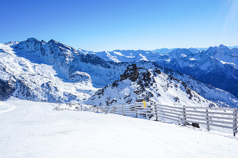 Breite, präparierte Skipiste im Vordergrund, abgegrenzt durch einen langen Holzzaun auf der rechten Seite. Dahinter erstrecken sich schroffe, schneebedeckte Bergformationen unter einem klaren, hellblauen Himmel bei Sonnenschein. Val Thorens Frankreich Schwarze Piste Orelle