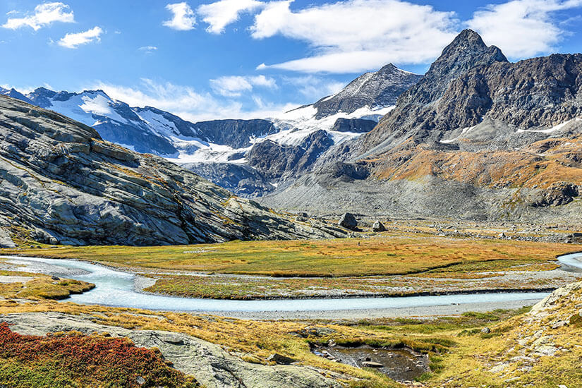 Ein geschwungener, hellblauer Bergfluss schlängelt sich durch ein hochalpines Tal im Vanoise-Nationalpark. Im Hintergrund ragen felsige, steile Berggipfel mit vereinzelten Schneefeldern und Gletscherresten unter blauem, leicht bewölktem Himmel empor. Val Thorens Frankreich Vanoise