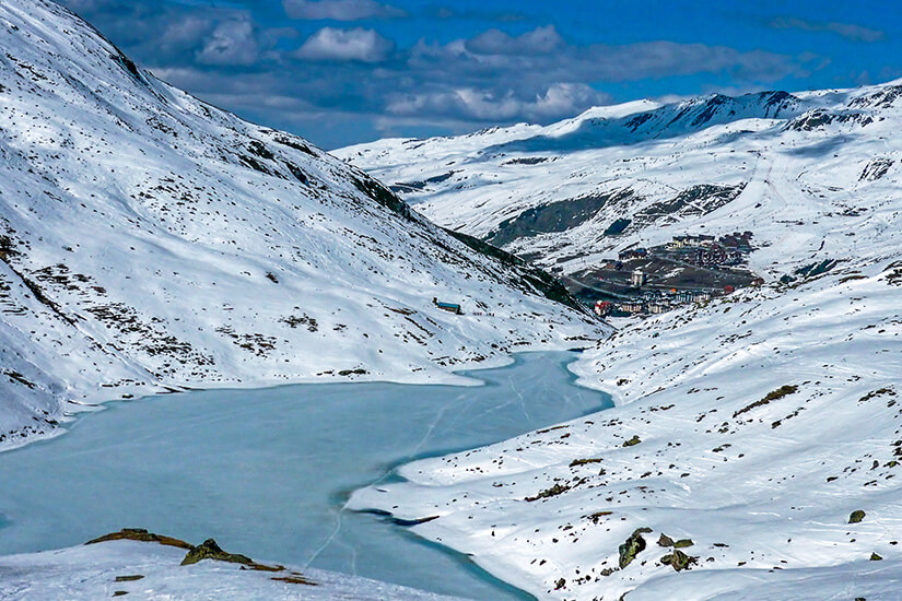Blick von oben auf den zugefrorenen, leicht blau schimmernden Lac du Lou, der eingebettet in einem verschneiten Tal liegt. Im Hintergrund sind schneebedeckte Berge und weiter entfernt der Ort Val Thorens zu sehen. Val Thorens Frankreich Lac du Lou