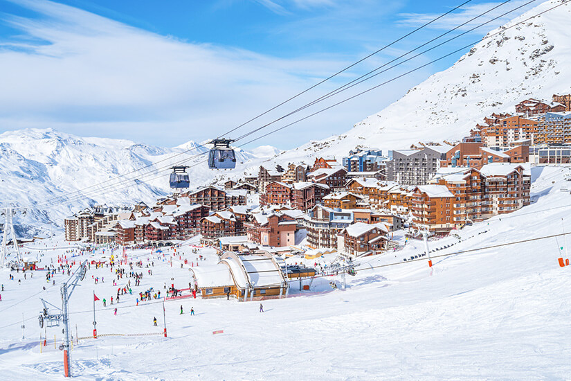 Panoramablick auf den verschneiten Wintersportort Val Thorens bei strahlendem Sonnenschein und blauem Himmel. Im Vordergrund Schweben moderne Gondeln an Seilen über die belebten, schneebedeckten Pisten vor der Kulisse alpiner Bauten und Berggipfel. Val Thorens Frankreich Val Thorens