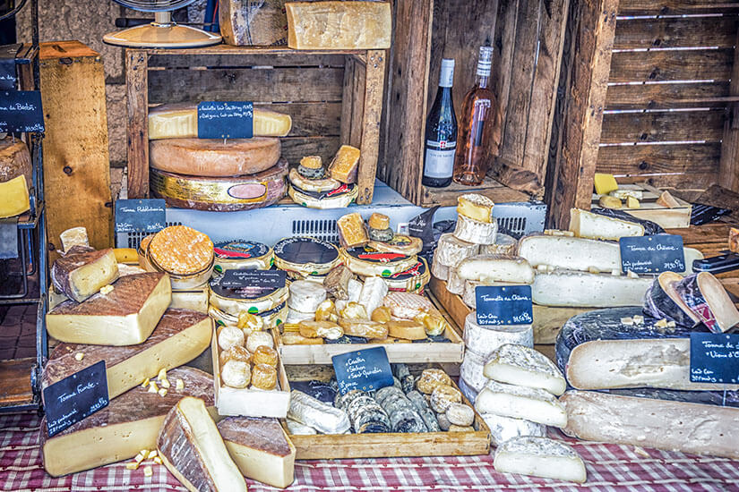 Ein reich gefüllter Marktstand mit zahlreichen traditionellen savoyischen Käsesorten, darunter große Käseräder, Ziegenkäselaibe und Schachteln mit Reblochon. Im Hintergrund stehen Holzkisten mit passenden Weinflaschen und Preisschildern. Val Thoresn Frankreich Kaese