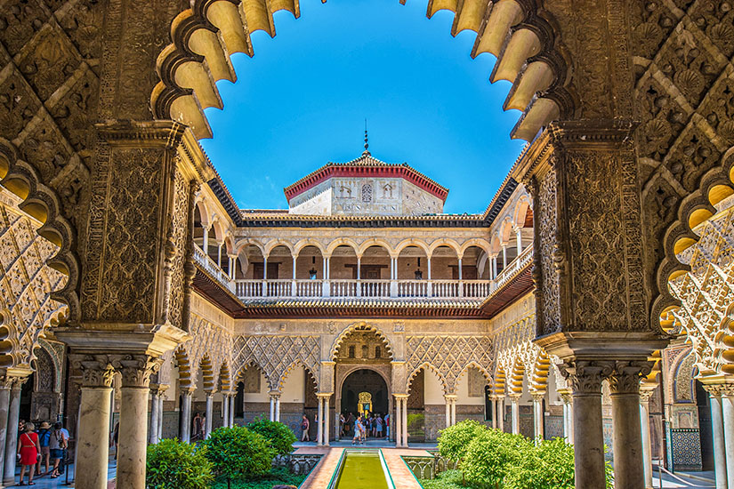 Durch einen kunstvoll verzierten Bogen im Mudéjar-Stil fällt der Blick auf den Patio de las Doncellas im Real Alcázar von Sevilla, dessen langes Wasserbecken von grünen Hecken und filigranen Arkaden gesäumt wird. Game of Thrones Drehorte Sevilla