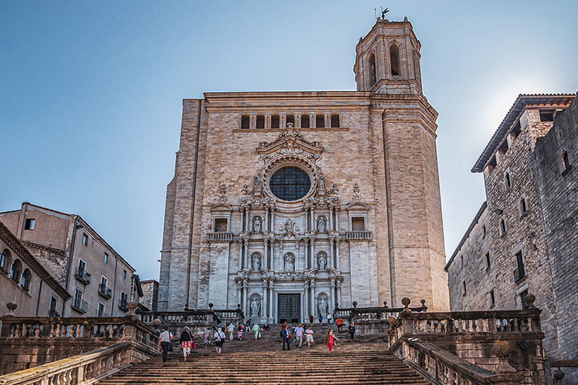 Über eine breite, monumentale Freitreppe erreicht man die Kathedrale von Girona, deren barocke Fassade mit großer Fensterrosette, Heiligenfiguren und hohem Glockenturm majestätisch in den Himmel ragt. Game of Thrones Drehorte Girona