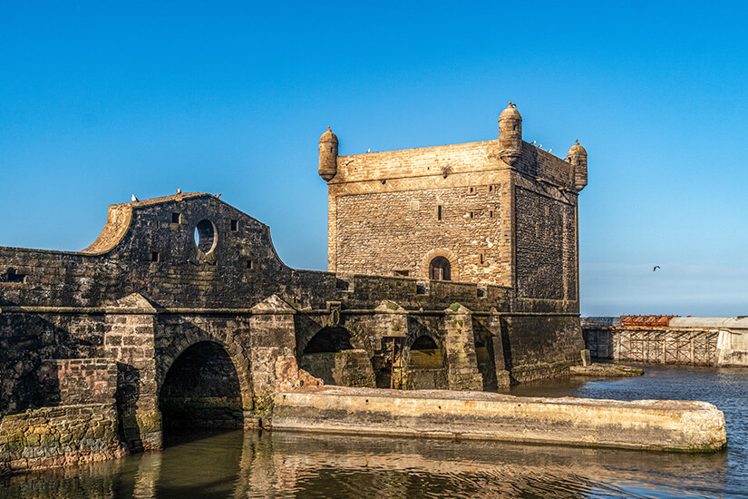 Direkt am Wasser erhebt sich der wuchtige Wehrturm der Skala du Port in Essaouira, dessen verwitterte Steinmauern mit kleinen Ecktürmchen sich im ruhigen Hafenbecken spiegeln. Game of Thrones Drehorte Essaouira