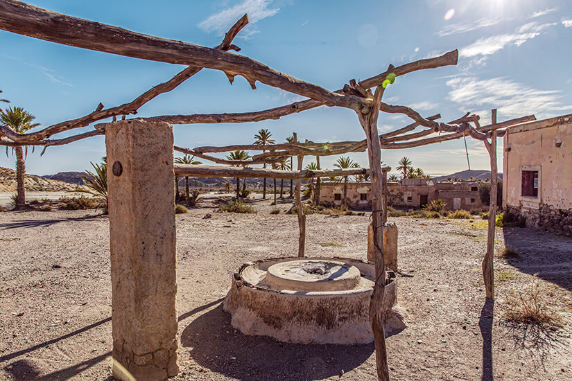 Unter einer rustikalen Pergola aus knorrigen Ästen steht ein alter, gemauerter Brunnen inmitten einer staubigen Wüstenlandschaft bei Almería. Game of Thrones Drehorte Almeria