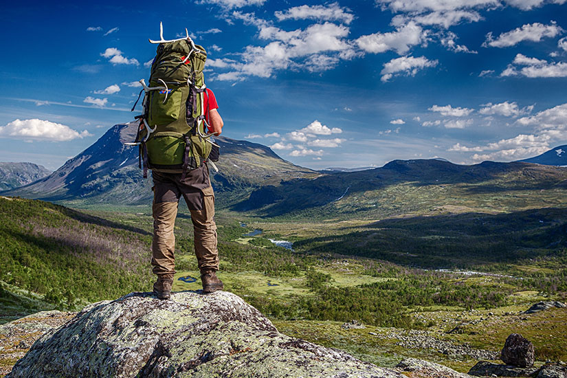 Auf einem flechtenbewachsenen Felsen steht ein Wanderer mit großem, grünem Trekkingrucksack, an dem ein Rentiergeweih befestigt ist, und blickt über die weite Fjelllandschaft. Packliste Finnland Ausblick