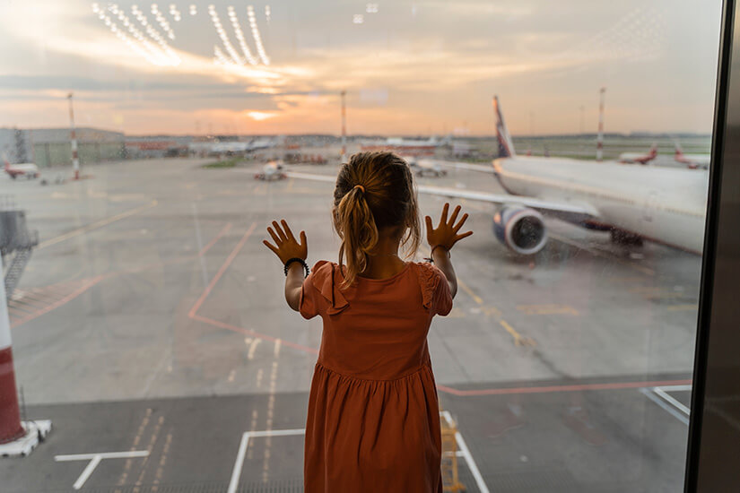 Mit beiden Händen an die große Glasscheibe gelehnt, schaut ein kleines Mädchen im rostroten Kleid gebannt auf das Vorfeld eines Flughafens hinaus. Co2 Rechner Reisen Flughafen