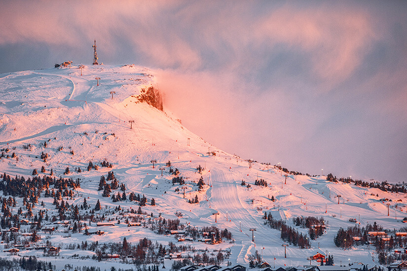 Ein schneebedeckter Bergkamm im Skigebiet Skeikampen, dessen Hänge und Lifte im rosafarbenen Licht des Sonnenuntergangs leuchten. Am Fuß des Berges liegen kleine Holzhütten verstreut zwischen verschneiten Bäumen. Skigebiete Norwegen Skeikampen